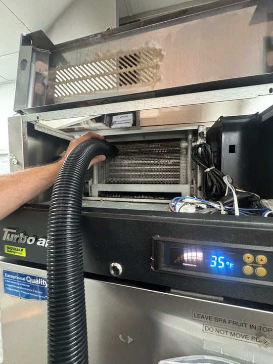 Technician vacuuming the condenser of a Turbo Air commercial refrigerator while the display reads 35 degrees Fahrenheit